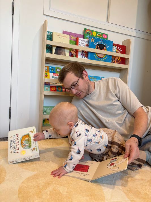 Man and child reading books together on a wooden floor with a bookshelf in the background.